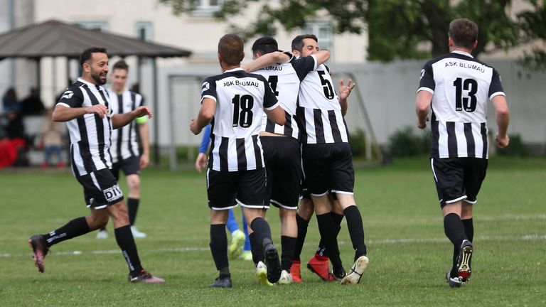 Fußballspieler feiern auf einem Feld, wobei sich zwei Spieler umarmen. Sie tragen schwarze und weiße Trikots mit Nummern wie 18, 4 und 5. Bäume und ein Gebäude sind im Hintergrund zu sehen.
