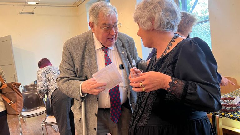 Ein älterer Mann in Anzug und Krawatte zeigt einer älteren Frau mit einem Getränk auf einer Zusammenkunft ein Dokument.