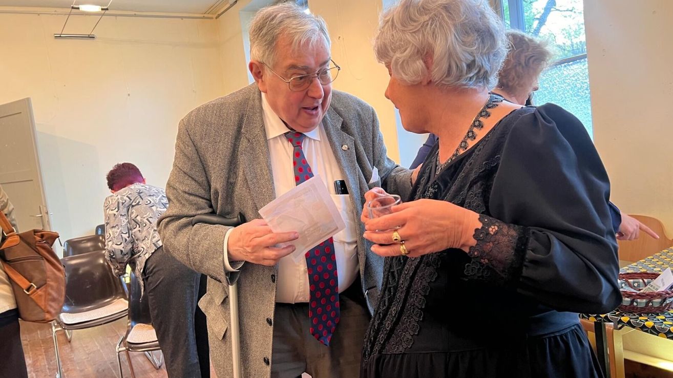 Ein älterer Mann in Anzug und Krawatte zeigt einer älteren Frau mit einem Getränk auf einer Zusammenkunft ein Dokument.