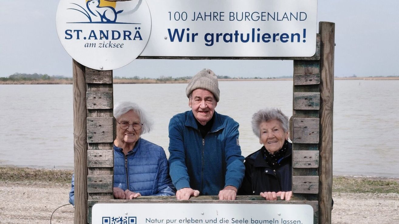 Drei ältere Menschen, lächelnd, stehen am See mit einem Schild, das '100 Jahre Burgenland. Wir gratulieren!' besagt. Die Frau links trägt eine Brille und eine blaue Jacke, der Mann in der Mitte eine blaue Jacke und eine Mütze, und die Frau rechts eine schwarze Jacke. Unten befindet sich ein QR-Code.