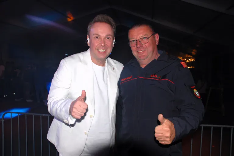 Zwei Männer posieren nachts für ein Foto, einer in einem weißen Anzug mit Daumen hoch und der andere in einem schwarzen Mantel mit rotem Saum.