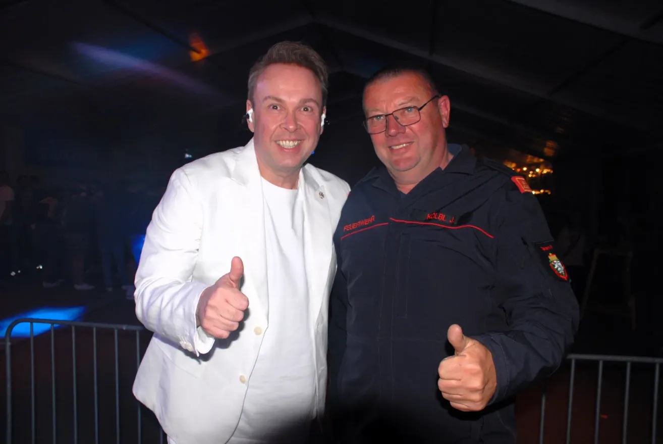 Zwei Männer posieren nachts für ein Foto, einer in einem weißen Anzug mit Daumen hoch und der andere in einem schwarzen Mantel mit rotem Saum.