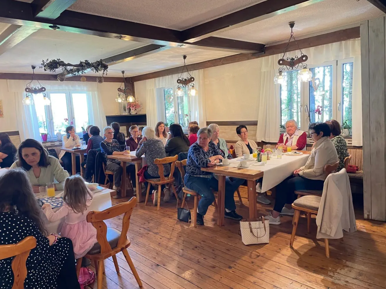 Eine Gruppe von Erwachsenen und Kindern sitzt an Tischen in einem Restaurant und isst Frühstück. Eine Frau hält ein Kind. Der Raum hat weiße Wände und Holzböden.