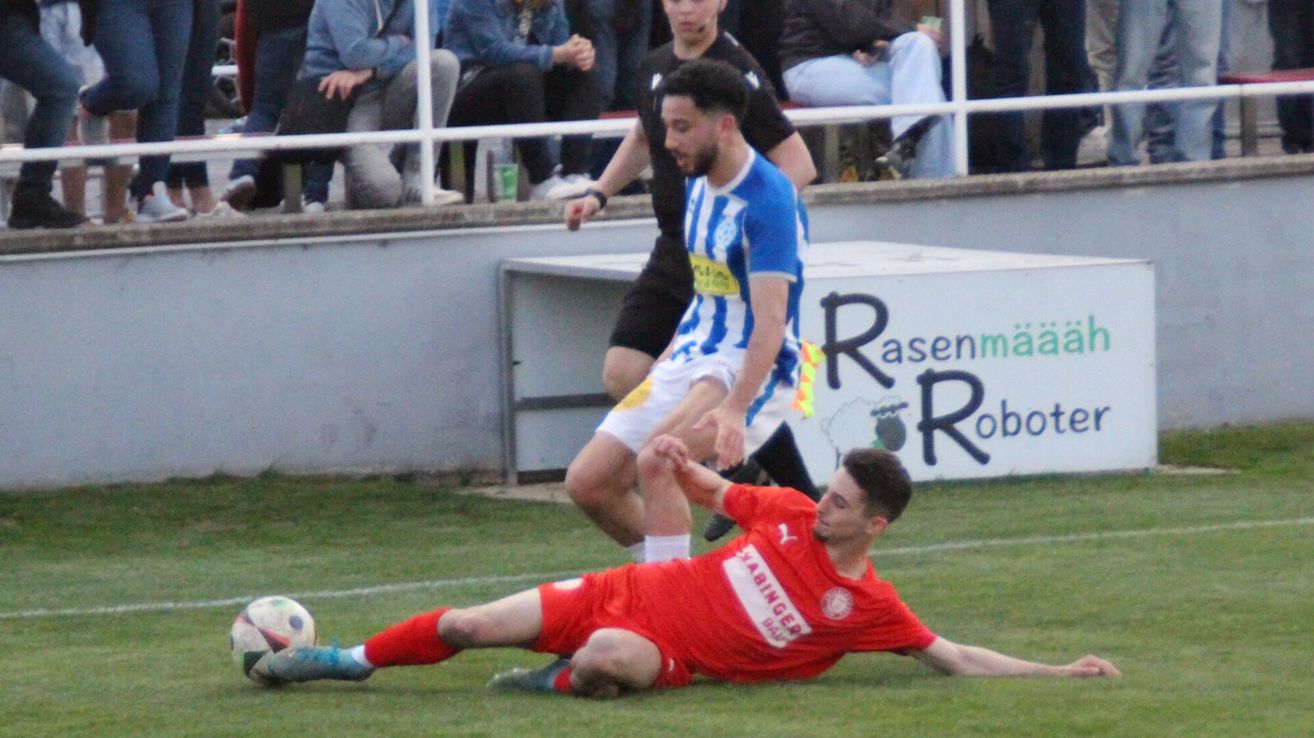 Fußballspieler bei einem Spiel, einer in Rot liegt auf dem Boden, während ein anderer in Blau verteidigt. Zuschauer beobachten von einer weißen Absperrung aus.