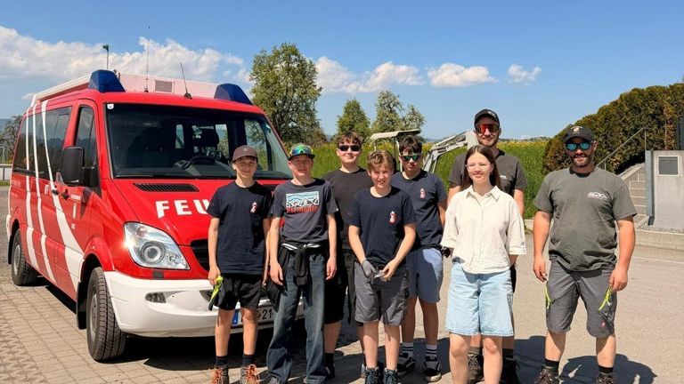 Eine Gruppe junger Leute steht vor einem roten Feuerwehrauto. Sie tragen Freizeitkleidung, einige haben eine Brille auf, und zwei halten Handschuhe. Ein Maschinenfahrzeug ist im Hintergrund zu sehen.