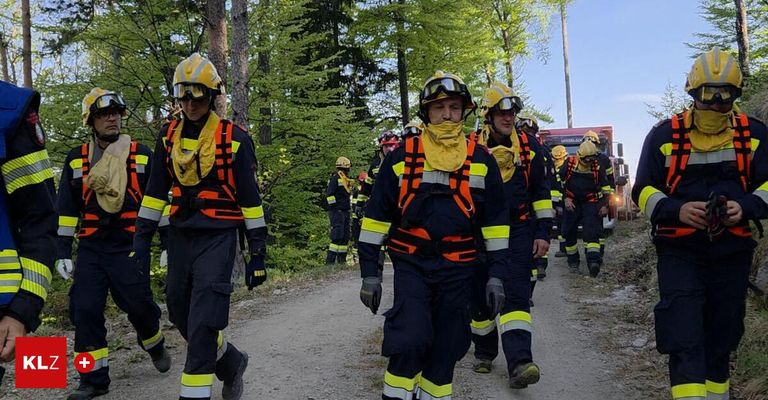 Eine Gruppe Feuerwehrleute geht in voller Ausrüstung auf einem unbefestigten Weg. Bäume und ein Feuerwehrauto sind im Hintergrund zu sehen.