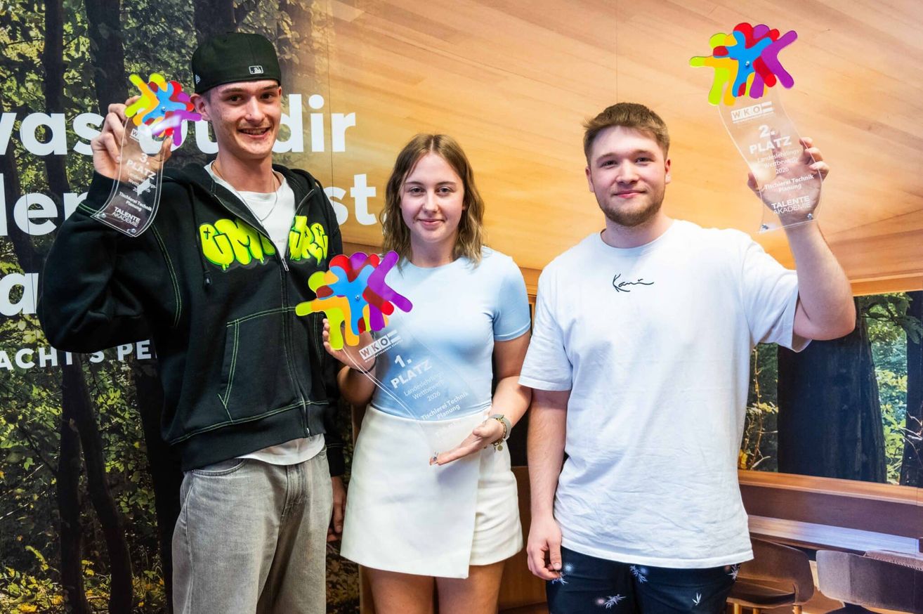 Drei junge Erwachsene posieren mit Trophäen für ein Foto. Die Frau hält einen bunten Preis. Hinter ihnen zeigt eine hölzerne Kulisse ein Eventlogo.