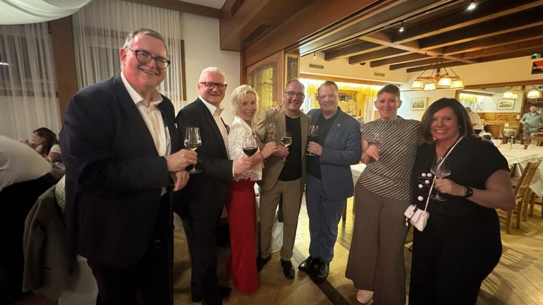 A group of six people are standing together, holding wine glasses, and smiling in a room.