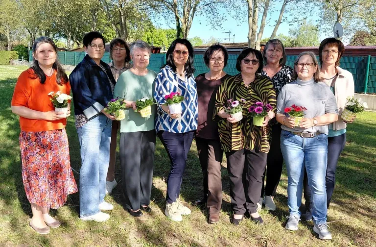 Neun Frauen stehen in einer Reihe in einem Park, jede mit einem Blumentopf. Sie sind lässig gekleidet und lächeln für die Kamera. Hinter ihnen steht ein grüner Zaun und Bäume.