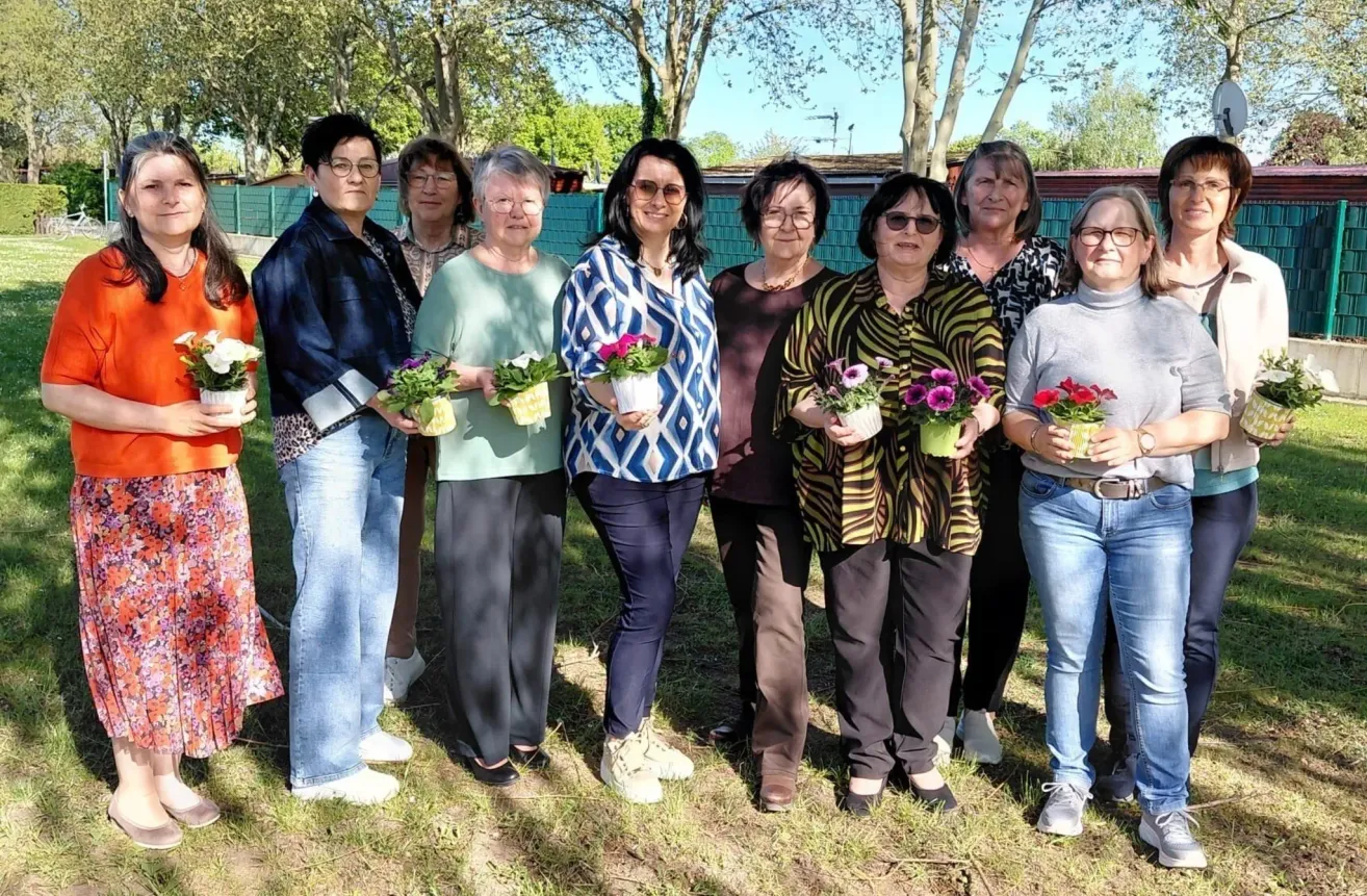 Neun Frauen stehen in einer Reihe in einem Park, jede mit einem Blumentopf. Sie sind lässig gekleidet und lächeln für die Kamera. Hinter ihnen steht ein grüner Zaun und Bäume.