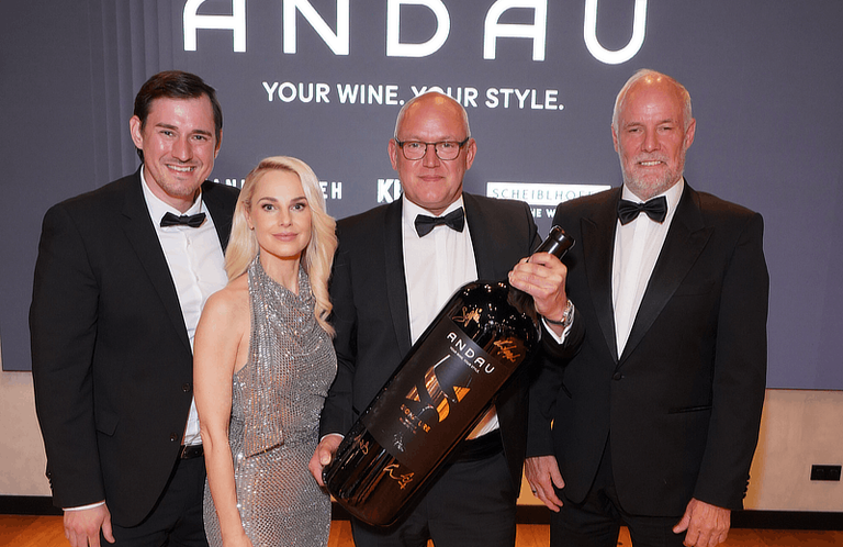 Vier Personen stehen in formeller Kleidung zusammen. Ein Mann hält eine große Flasche Wein. Hinter ihnen steht ein Banner mit der Aufschrift 'Ihr Wein. Ihr Stil.'