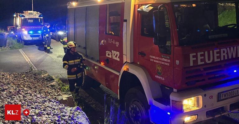 A firefighter stands next to a red fire truck at night. Several people are nearby, and lights are on.