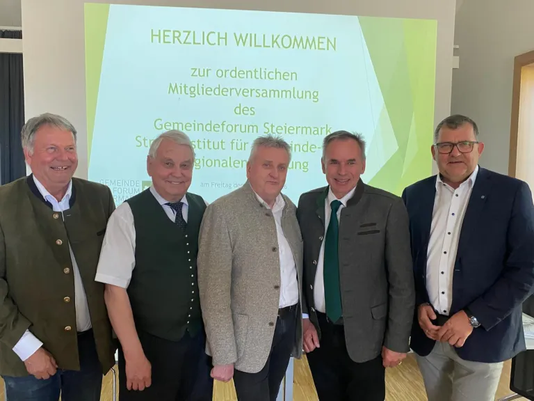 Fünf Männer stehen vor einer Projektionsleinwand, auf der Herzlich Willkommen zur ordentlichen Mitgliederversammlung des Gemeindeforum Steiermark steht. Sie tragen formelle Kleidung, und drei von ihnen lächeln.