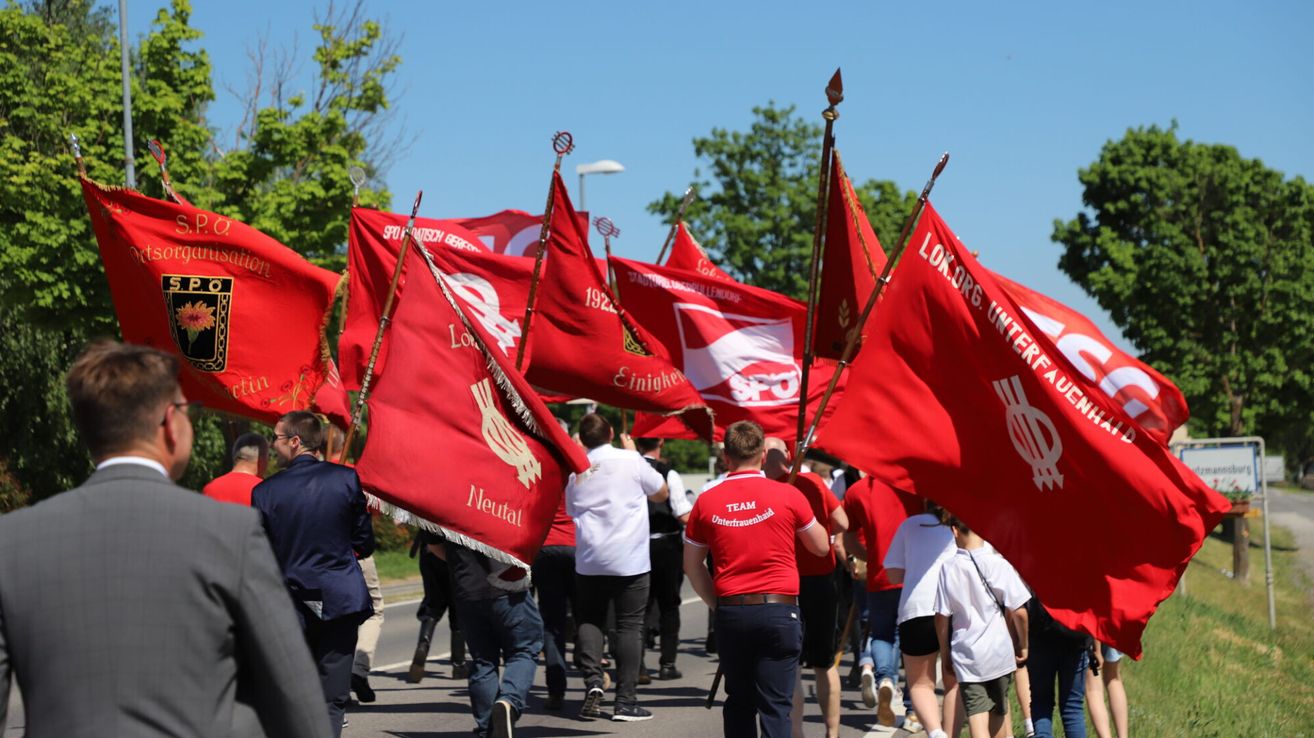 Eine Gruppe von Personen marschiert auf einer Straße und hält rote Fahnen mit verschiedenen Designs und Texten in der Hand, unter einem klaren blauen Himmel. Bäume säumen die Straßenseiten.