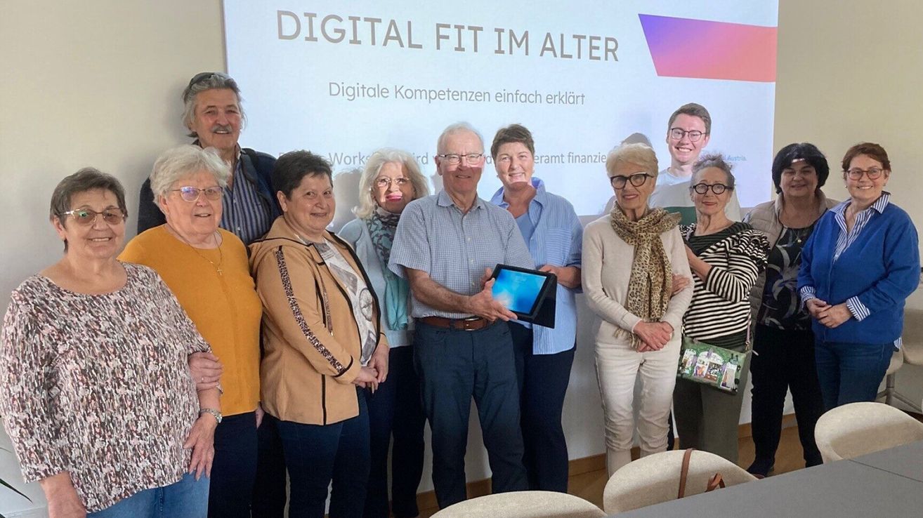 Eine Gruppe älterer Menschen steht in einem Klassenzimmer. Ein Mann hält ein Tablet und eine andere Person steht neben ihm. Ein Whiteboard ist vor ihnen.