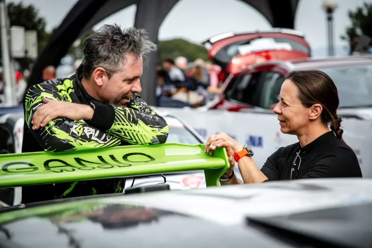 Ein Mann und eine Frau lächeln und betrachten ein grünes Objekt an einem Auto auf einem Parkplatz. Der Mann trägt einen schwarzen und grünen Rennanzug.
