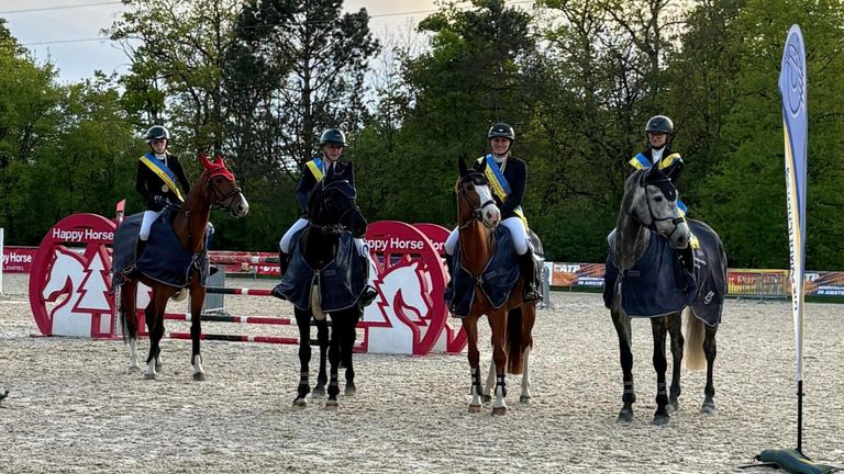 Three riders on horses stand in a field. Each rider wears a helmet, and the horses have blue covers. Behind them is a red sign with a white horse. Trees are in the background.