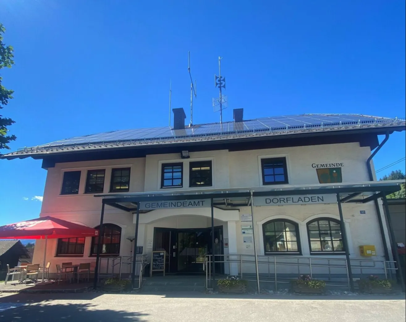 Das Gebäude verfügt über Solarmodule auf dem Dach, ein Schild mit der Aufschrift Gemeindeamt Dorfladen und mehrere Fenster. Draußen befindet sich ein roter Regenschirm und Stühle.
