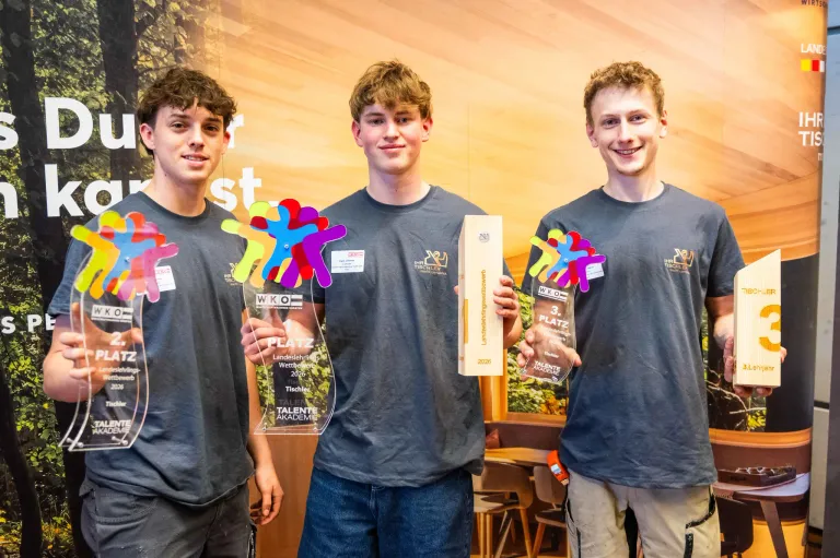 Drei junge Männer stehen zusammen und halten Trophäen. Sie tragen passende graue T-Shirts mit Logos. Der Mann links hält eine bunte Papierfigur, der in der Mitte eine hölzerne Box und der rechts eine Trophäe mit '3. PLATZ' darauf. Hinter ihnen ist eine Wand mit einem warmen Hintergrund und etwas Text.