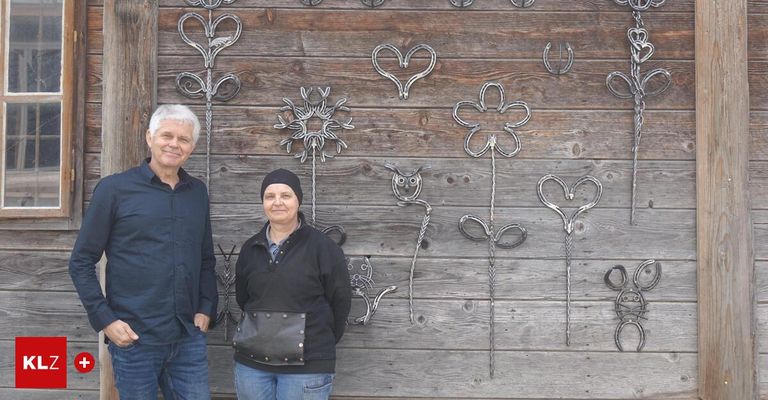 Zwei Personen stehen neben einer Holzwand, die mit verschiedenen metallischen Blumendesigns wie Herzen und Schneeflocken verziert ist.