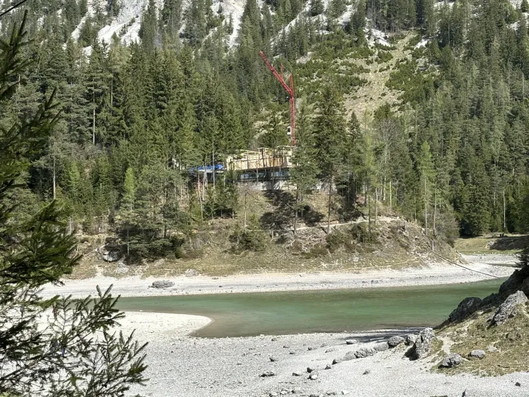 Ein großer Kran steht auf einem Hang mit einem im Bau befindlichen Gebäude, umgeben von einem üppigen Wald und einem Fluss unten.