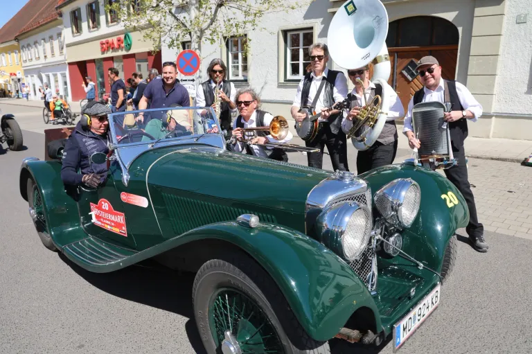 Eine Gruppe von Musikern mit Sonnenbrillen und Hüten spielt verschiedene Instrumente vor einem grünen Oldtimer, wahrscheinlich bei einem Umzug.