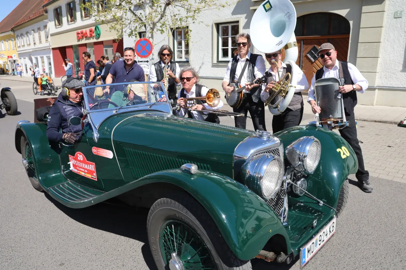 Eine Gruppe von Musikern mit Sonnenbrillen und Hüten spielt verschiedene Instrumente vor einem grünen Oldtimer, wahrscheinlich bei einem Umzug.