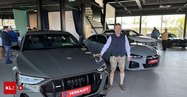 Ein Mann steht neben einem grauen Audi mit einem 'VERKAUFT'-Aufkleber in einer modernen Autowerkstatt mit anderen Autos im Hintergrund.