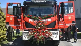 Ein roter Feuerwehrwagen namens Takern II ist geparkt, mit offenen Türen. Ein Blumenkranz ziert die Vorderseite. Zwei Personen, eine in einer reflektierenden Weste, stehen in der Nähe.