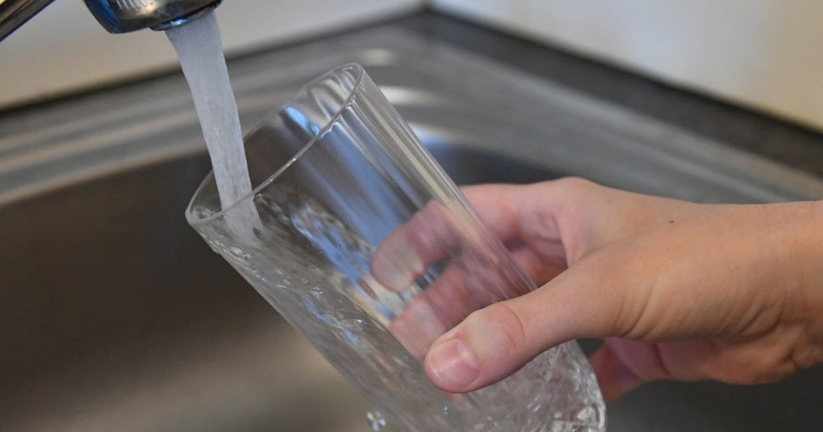 Eine Hand füllt ein Glas mit Wasser aus einem Wasserhahn in einer Küche. Das Glas ist durchsichtig und der Wasserhahn ist silber.