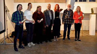 Eine Gruppe von Personen steht zusammen und hält Schriftrollen in den Händen, vermutlich bei einer Preisverleihung. Sie tragen verschiedene Kleidungsstücke und scheinen für ein Foto zu lächeln.