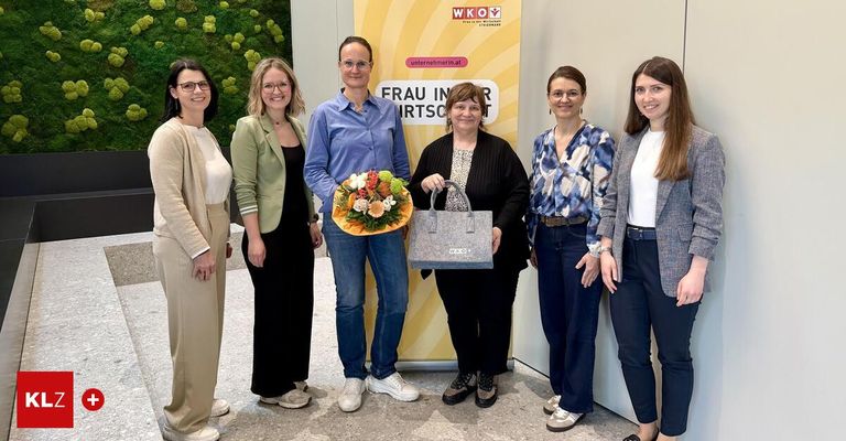 Sechs Frauen posieren vor einem Banner mit dem Text 'Frau in der Wirtschaft', halten einen Blumenstrauß und eine Tasche in der Hand, wahrscheinlich bei einer Veranstaltung.
