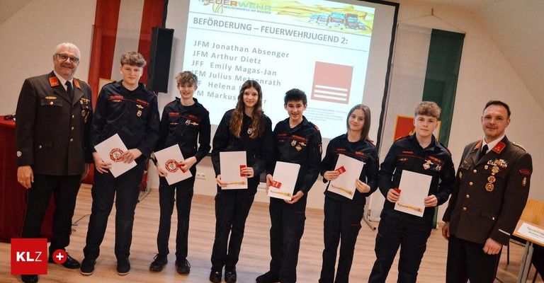 Seven young people dressed in black uniforms stand before a projector screen with various names on it, holding papers. They seem to be posing for a photograph.