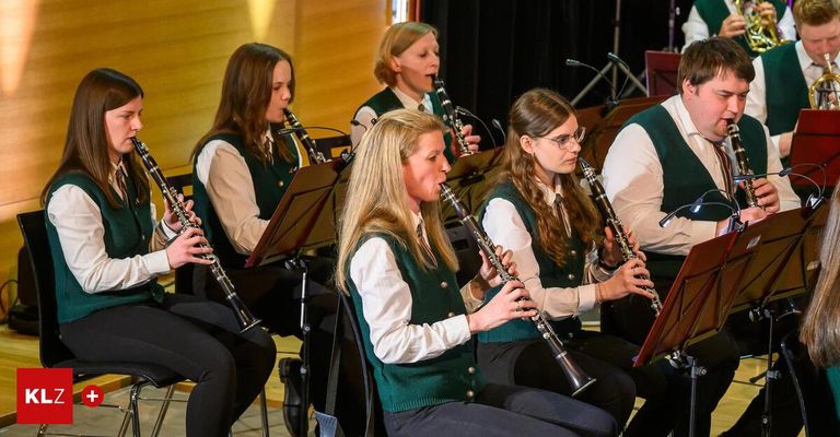 Fünf Musiker sitzen in einer Reihe und spielen Klarinette. Eine Frau in der Mitte trägt eine Brille und eine grüne Weste. Eine andere trägt ein weißes Hemd. Sie spielen wahrscheinlich in einer Schule oder einem Musiksaal.