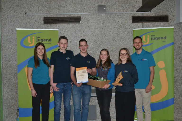 Sechs Personen stehen lächelnd vor einer Wand. Drei Männer und drei Frauen. Zwei Frauen halten Auszeichnungen. Der Mann in der Mitte hält eine Schachtel mit Süßigkeiten und ein Zertifikat. Hinter ihnen befinden sich zwei Banner mit dem Text Jugend.