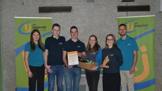 Sechs Personen stehen lächelnd vor einer Wand. Drei Männer und drei Frauen. Zwei Frauen halten Auszeichnungen. Der Mann in der Mitte hält eine Schachtel mit Süßigkeiten und ein Zertifikat. Hinter ihnen befinden sich zwei Banner mit dem Text Jugend.