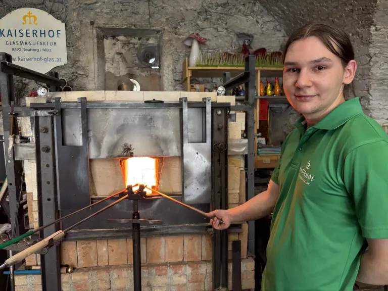 Ein Mann steht vor einem Ofen, hält Werkzeuge und lächelt. Hinter ihm befindet sich ein Backofen aus Ziegeln mit offener Tür, einer Steinmauer und verschiedenen Gegenständen auf einem Regal.