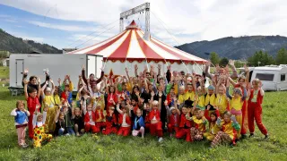 Eine Gruppe von Kindern in bunten Kostümen posiert für ein Foto vor einem großen Zirkuszelt. Im Hintergrund sind Berge zu sehen.