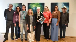Sieben Frauen stehen vor einer weißen Wand, drei Gemälde dahinter. Eine Frau hält ein Buch mit dem Titel 'Die Gesse der Frauen'.