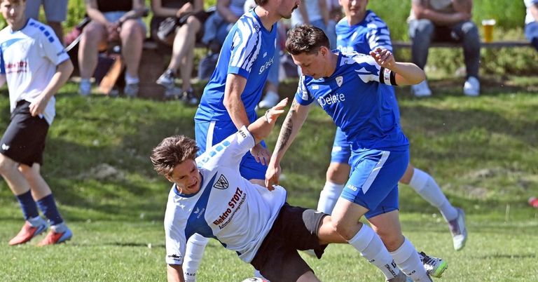Ein Fußballspiel mit Spielern in blau-weißen Trikots, ein Spieler in Weiß fällt zu Boden, ein anderer verteidigt ihn.