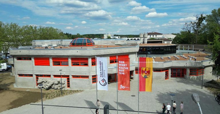 Luftaufnahme eines modernen Gebäudes mit Fahnen. Es ist das Frank Hofmann Kulturzentrum. Leute laufen auf dem Bürgersteig.