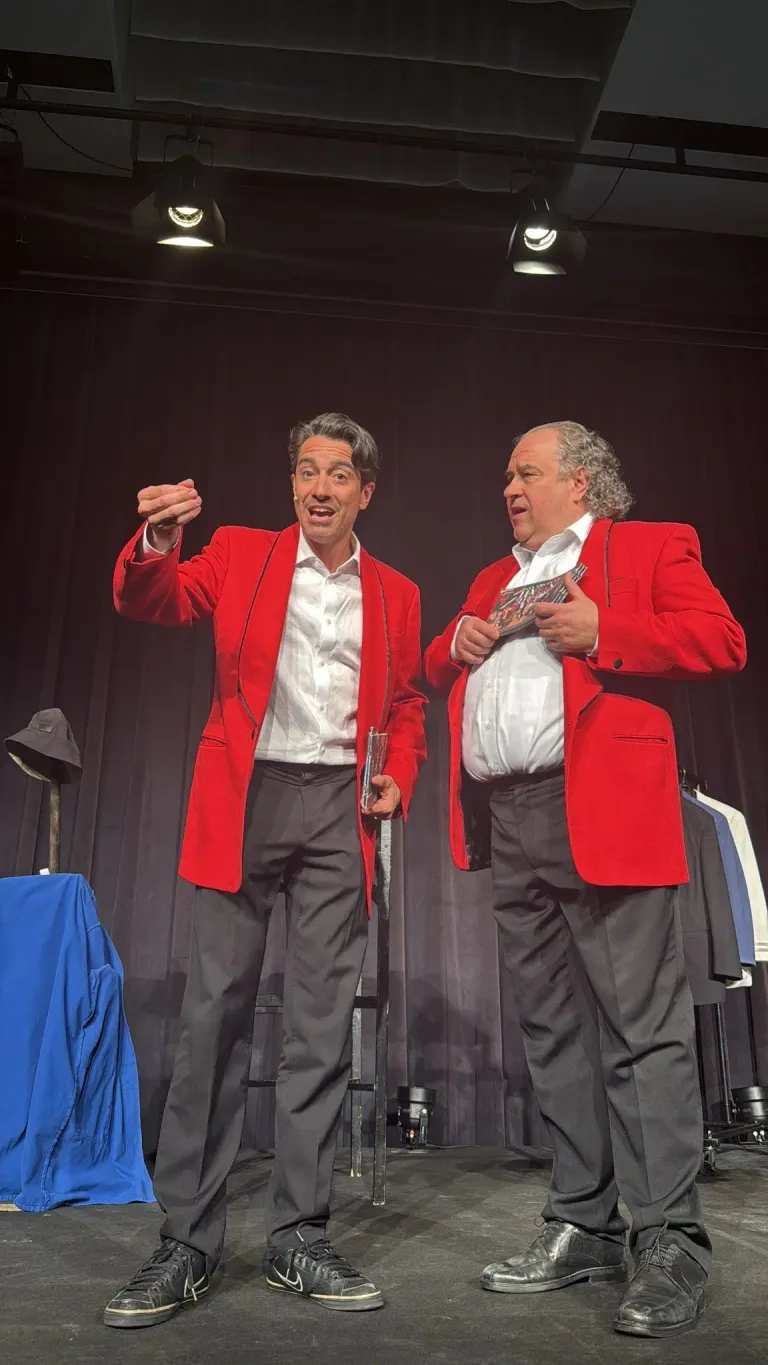 Two men in red blazers and white shirts on stage. One holds a glass, the other a folded magazine. Behind them, a black curtain with a blue cloth and a hat.