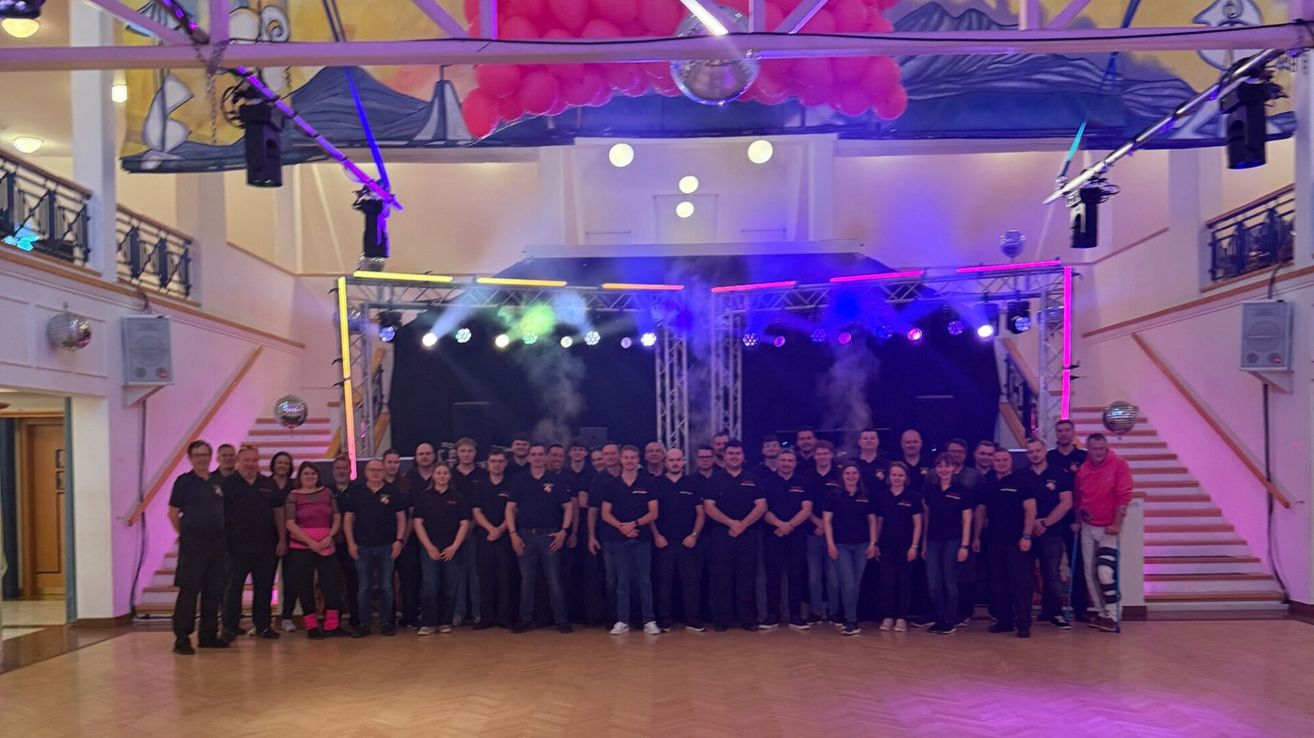 Eine Gruppe von Menschen, möglicherweise ein Team, steht auf einer Bühne mit einem Discoball, Luftballons und Bühnenbeleuchtung. Einige tragen schwarze T-Shirts mit Logos.
