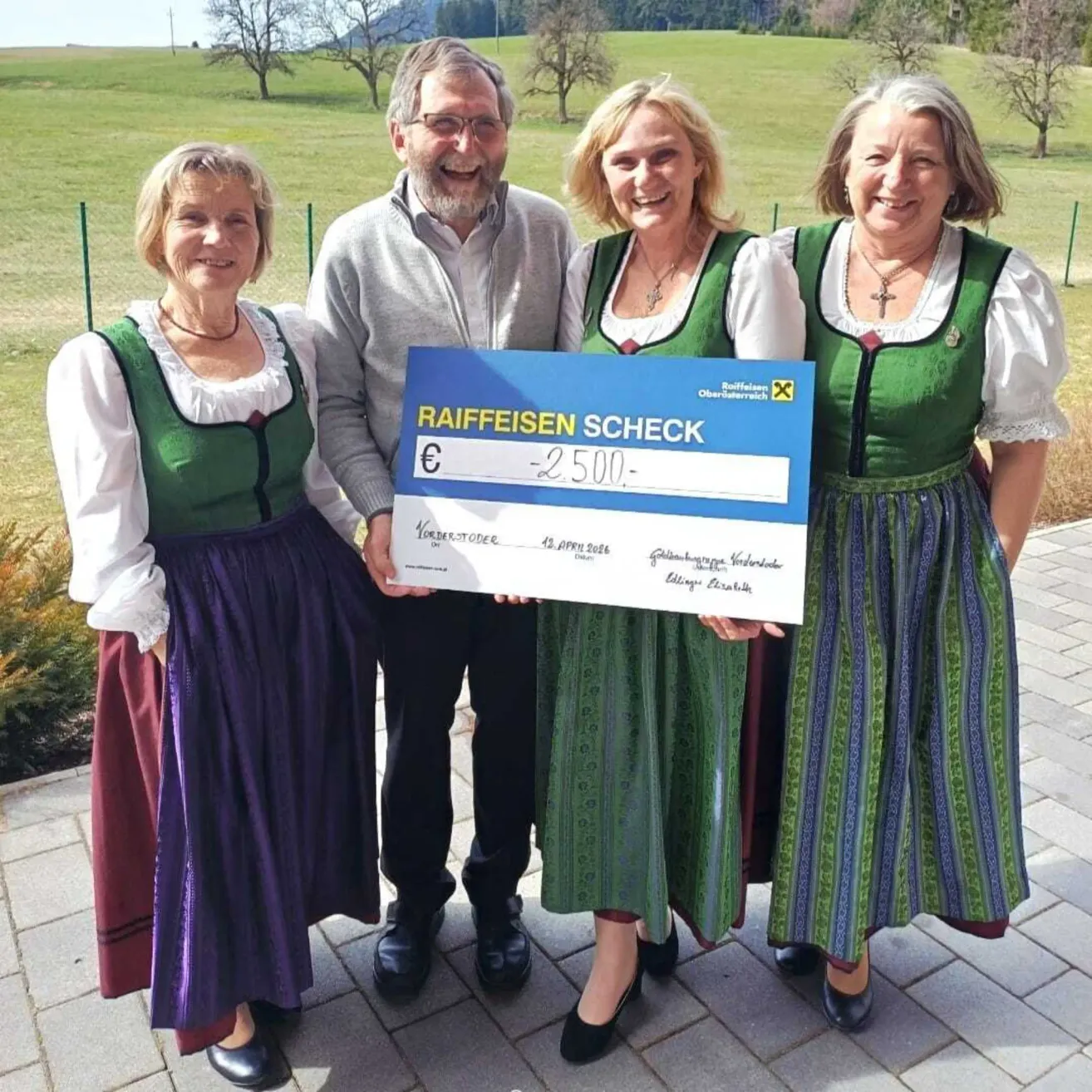 Vier Personen, zwei Männer und zwei Frauen, stehen in einem Feld, lächeln und halten einen großen Scheck in der Hand. Sie tragen alle traditionelle grüne Kleider. Hinter ihnen ist ein Feld und Bäume zu sehen.