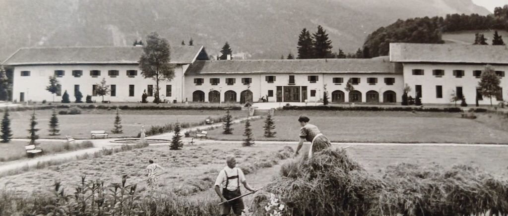 Ein Schwarz-Weiß-Foto zeigt ein großes Gebäude mit einem Garten davor. Zwei Personen sind damit beschäftigt, im Garten Ernte einzusammeln.