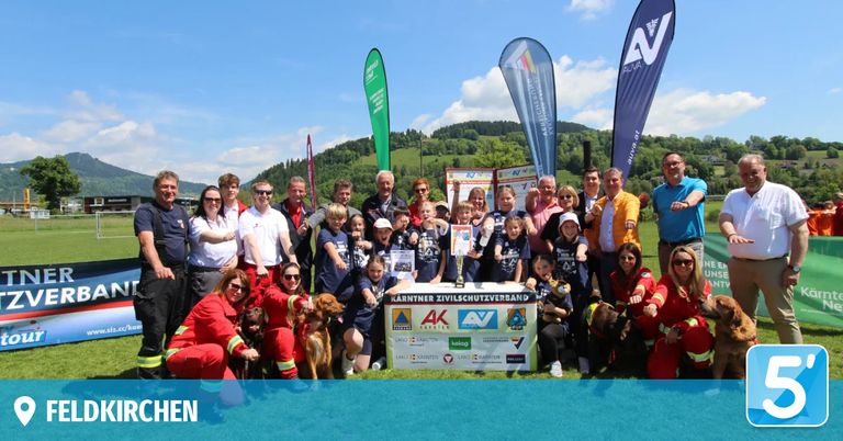 Eine Gruppe von Menschen und Hunden posiert mit einer Trophäe und Bannern für ein Foto. Einige tragen rote Uniformen. Der Hintergrund zeigt einen Berg mit Bäumen.