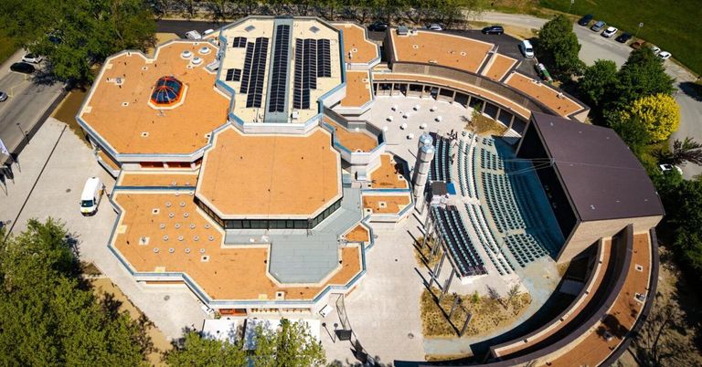 Ein Blick von oben auf ein architektonisches Gebäude mit Solarpaneelen auf dem Dach und einem Sitzbereich darunter. Die Struktur ist von Grün und Bäumen umgeben.