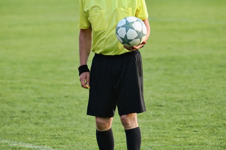 A soccer referee holds a soccer ball, standing on a grassy field. He wears a yellow shirt, black shorts, and black socks.