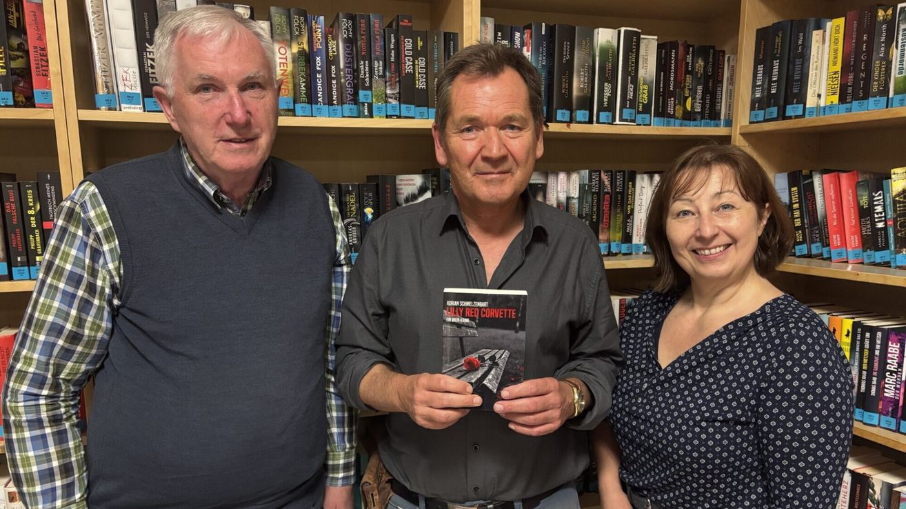 Drei Personen stehen vor einem Bücherregal voller Bücher. Der Mann in der Mitte hält ein Buch mit dem Titel 'Lilly Red Corvette.' Der Mann links trägt ein kariertes Hemd und die Frau rechts ein gepunktetes Kleid.