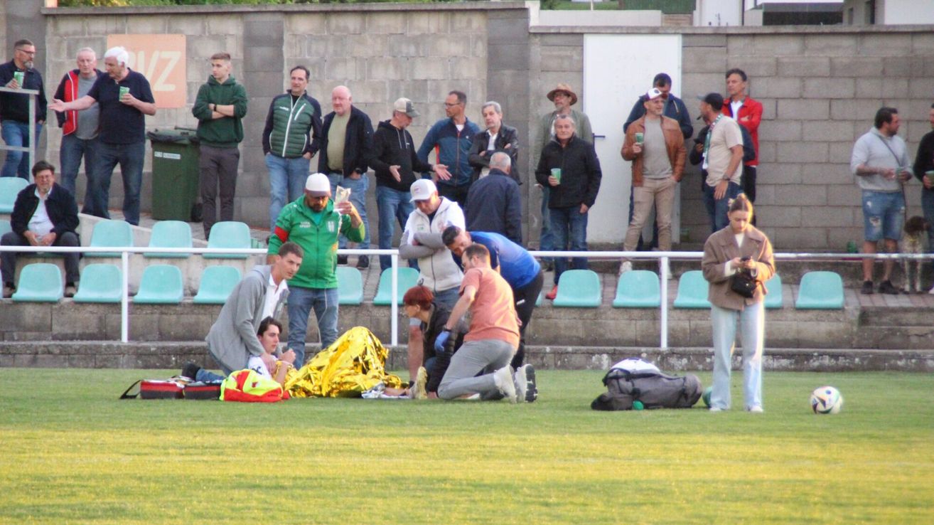 Ein Mann auf einem Fußballfeld wird von medizinischem Personal versorgt. Zuschauer stehen hinter einer Barriere, einige mit Getränken.
